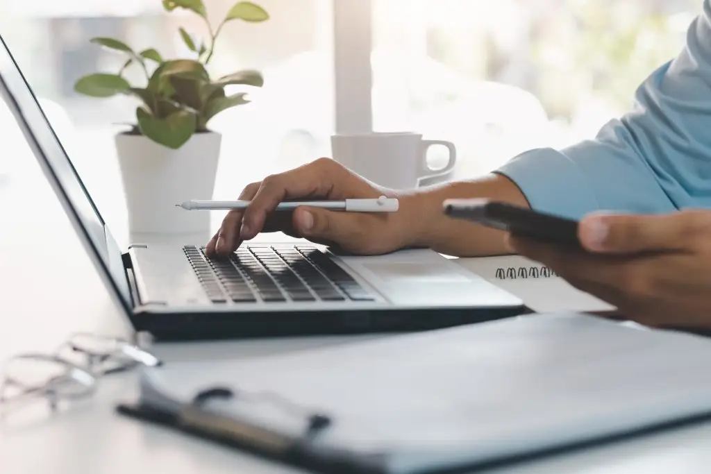 Hand mit Stift und Smartphone vor Laptop bei der Arbeit im Büro
